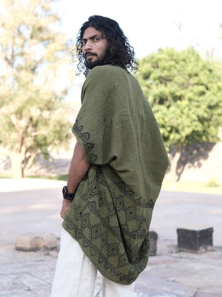 A man with curly hair and a beard stands outdoors, looking over his shoulder. He wears the green hand block-printed Cocoon Shrug over light pants, with trees and stone structures in the background.