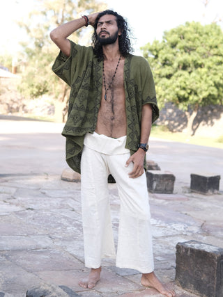 A barefoot man with long curly hair stands outdoors on stone pavement, wearing loose white cotton pants, a Cocoon Shrug layered over a beaded necklace and watch. He gazes upward with one hand behind his head.