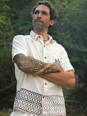 A man with curly hair and a patterned tattoo on his arm stands outdoors, arms crossed, thoughtfully gazing into the distance while wearing the Vairagya Shirt—a short-sleeved cream shirt crafted from organic cotton and adorned with decorative designs—perfectly embodying a sense of conscious fashion.