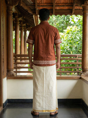 A man stands on a wooden veranda, facing away. He wears the Adhir Shirt—a brown, handcrafted-inspired cotton shirt with patterns—paired with a traditional white dhoti featuring a gold border. Lush greenery can be seen in the background, lending an ethnic contemporary charm to the scene.