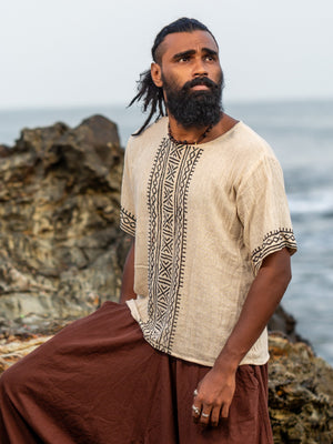 Model wearing the cream tribal block print t-shirt with a brown dhoti, standing against rocks by the sea, showing the hand block printed black geometric tribal motif down the centre front and the matching geometric sleeve trim on this unisex cotton tee.