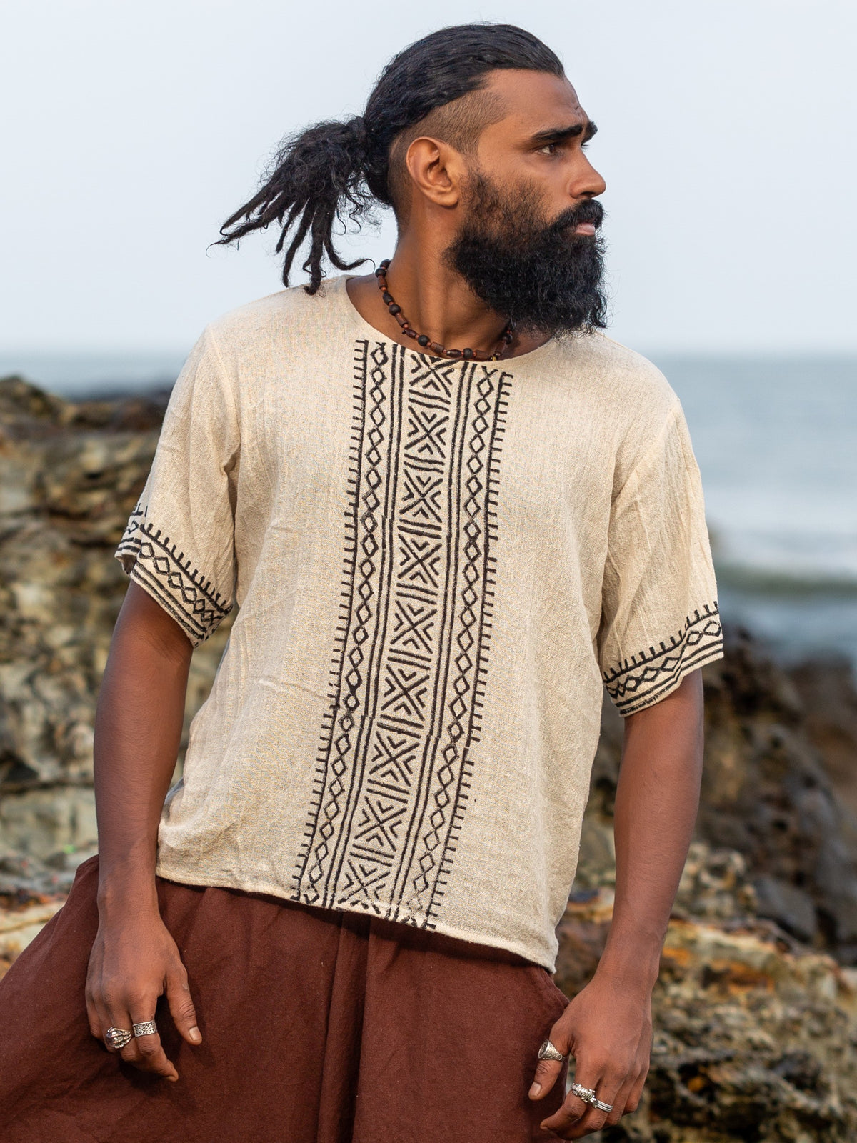 Front view of the model on a rocky shore in the cream tribal block print t-shirt and brown dhoti, showing the black hand block printed geometric tribal motif down the centre front and the matching geometric band around the short sleeves.