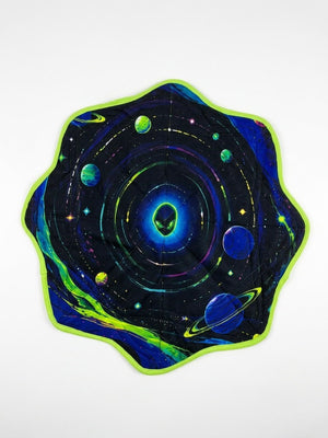 The Astral Dapo is a round, wavy-edged mat with a green border, showcasing colorful planets, stars, and cosmic planetary print patterns on a dark background that evoke a vibrant outer space scene. This quilted festival accessory infuses any gathering with interstellar energy.