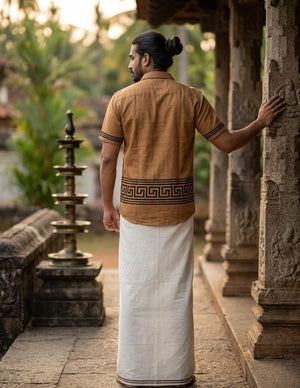 A man with long hair tied back stands outdoors near a stone pillar, wearing the Muraan Shirt—a relaxed fit, handblock printed brown shirt with black patterns—and a white traditional dhoti, facing away from the camera amid lush greenery.