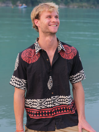 A smiling man with blonde hair wearing a Belgium Shirt featuring red and white patterns stands by a serene blue-green body of water.