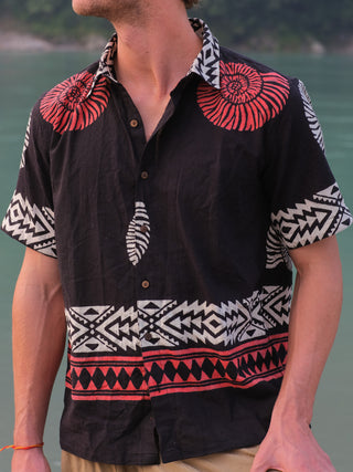 A man stands near water wearing the Belgium Shirt, a black short-sleeve top with bold red and white geometric and shell patterns inspired by Rajasthani handblock prints; his face is partially out of frame for a bohemian vibe.