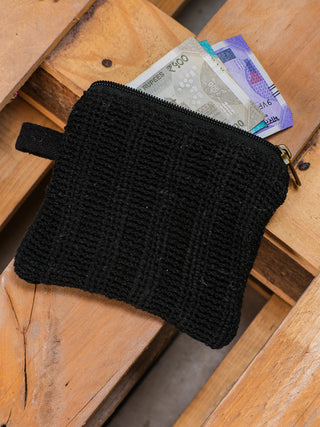 The sleek Upcycled Coin Pouch, made from black fabric and recycled materials, sits on a wooden surface with its zipper securing several paper currency notes peeking out.