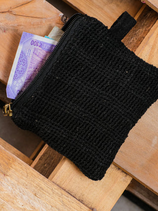 A partially open Upcycled Coin Pouch, crafted from repurposed materials, reveals purple currency notes inside. This versatile black fabric pouch with a zipper rests elegantly on a wooden surface.