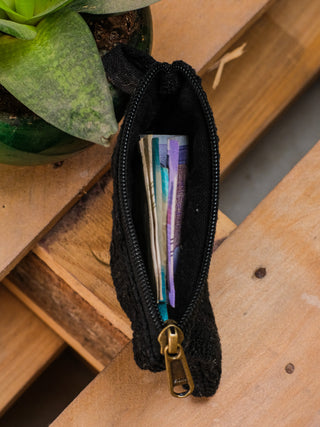 The Upcycled Coin Pouch, made from recycled materials, lies on a wooden surface with folded cash inside. A green potted succulent plant nearby enhances the natural vibe of the setting.