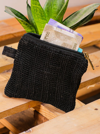 The Upcycled Coin Pouch, in black, rests on a wooden surface filled with currency notes. Its compact design is crafted from recycled materials, complemented by a backdrop of green leaves.