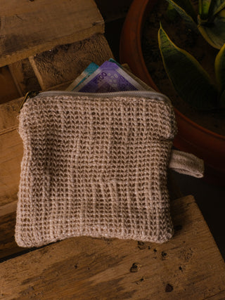 An Upcycled Coin Pouch in knitted beige rests on a wooden surface with its zipper partly open, showing colorful currency notes. Made from recycled materials, it sits near a vibrant potted plant.