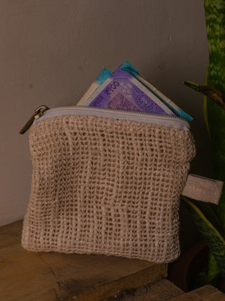 The Upcycled Coin Pouch, a beige woven accessory made from recycled materials, is partially open on a wooden surface, revealing colorful currency notes. A green plant peeks in from the right against a neutral backdrop.