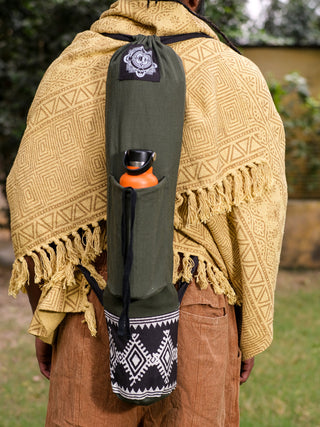 Wearing a yellow patterned shawl and brown pants, a person carries an olive green and black geometric Yoga Mat Bag with a side pocket for an orange water bottle, contrasting beautifully with the lush greenery and vibrant yellow wall.