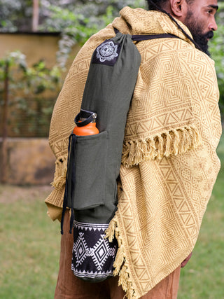 A bearded person in a patterned yellow shawl carries the Yoga Mat Bag, which features a tribal block print, green fabric, an orange bottle in its side pocket, and a black-and-white base. Theyre standing on grass with blurred greenery in the background.