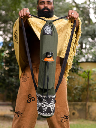 A person showcases the multi-functional Yoga Mat Bag, featuring a tribal block print in olive green. The bags black strap complements their outfit, which includes a yellow shawl and brown pants with symbolic designs. An orange water bottle peeks out as they stand against trees and a fence.