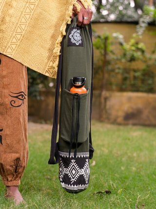 Standing on grass, a person cradles a Yoga Mat Bag featuring a tribal block print. A water bottle peeks from its pocket. Theyre dressed in a mustard yellow shawl and brown pants with black symbols, accentuating the bags multi-functional design.
