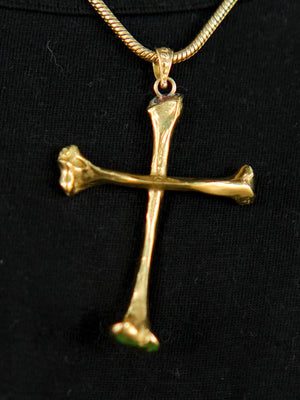 The Eternal Relic Necklace in gold features a pendant of two bone-like pieces, displayed on black fabric—a striking choice for gothic jewelry enthusiasts.