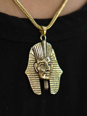Sure! Here’s a rewritten version using the product name: The Pharaoh Necklace features a gold pendant shaped like an ancient Egyptian pharaoh’s head, adorned with intricate lines and a majestic headdress. Worn over a black shirt, this piece is a striking tribute to traditional Egyptian jewelry.