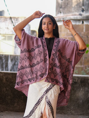 A woman stands outdoors in a loose, hand block-printed Cocoon Shrug with black geometric patterns over a fringed cream skirt, raising her arms slightly and looking confidently at the camera.