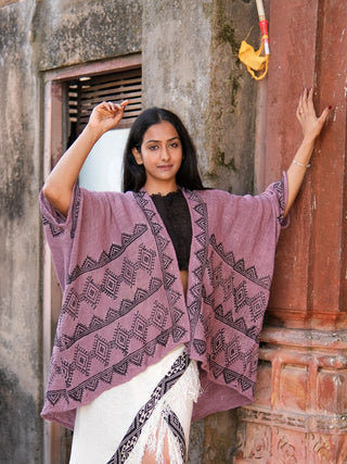 A woman with long dark hair stands by a red pillar and weathered wall, wearing the Cocoon Shrug in mauve over a black top and white fringed skirt, one hand raised to rest against the wall.