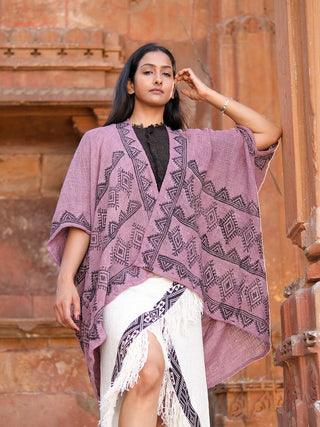 A woman stands confidently against a rustic stone backdrop, wearing the Cocoon Shrug in dusky pink over a fringed white skirt and black top. She has long dark hair and a calm, self-assured expression.