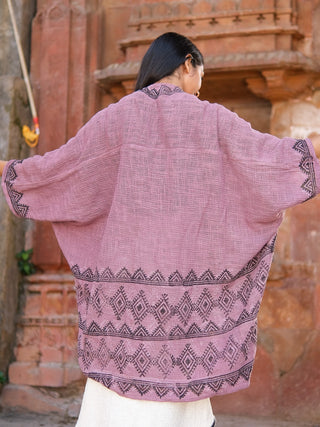 A person stands with their back to the camera, arms outstretched, wearing a mauve Cocoon Shrug made of cotton with black geometric patterns on the hem and sleeves. An ornate stone structure is visible in the background.