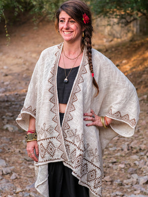 A smiling woman with long brown hair in a side braid stands outdoors on a dirt path, accessorized with jewelry and a red flower, wearing the Natural Miraya Shrug over a black top and pants—a truly stylish look.