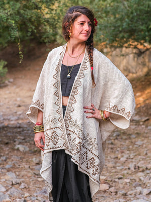 A woman with long braided hair stands outdoors amid greenery and rocks, wearing the beige patterned Miraya Shrug over a black top and skirt, accessorized with bracelets, rings, and a necklace.
