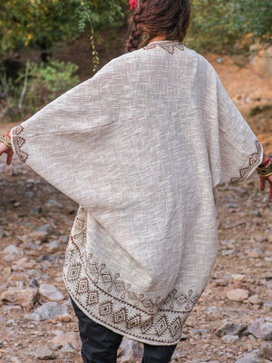 A person stands outdoors with their back to the camera, wearing a cream Miraya Shrug accented by brown geometric embroidery. Their dark hair contrasts with the natural rocks and greenery, highlighting this stylish accessory.