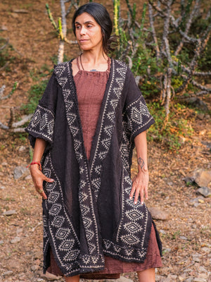 Front view of the model in the black knit kimono with the white block printed geometric border, showing the wide kimono sleeve and the long fringed hem over a dusty rose dress, in a desert cactus garden.