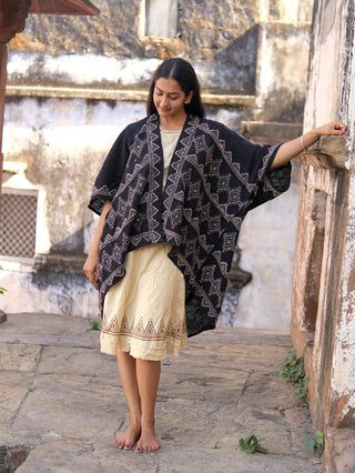 A woman stands barefoot on a stone path, wearing a cream dress with a hand block-printed hem and the Cocoon Shrug—an airy, black patterned cotton layer. She smiles gently, looking down, one arm resting on a weathered wall.
