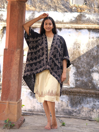 A woman stands barefoot outdoors by a weathered stone wall, wearing a cream dress with decorative trim and the hand block-printed cotton Cocoon Shrug.