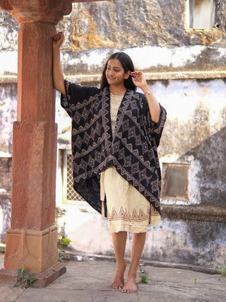 A woman stands barefoot on stone, leaning against a carved pillar. She wears a cream cotton dress paired with the Cocoon Shrug and smiles slightly, with a textured rustic wall in the background.