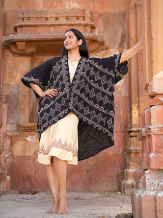 A woman poses confidently on stone steps against an ornate building, smiling upward. She wears a cream hand block-printed geometric dress with the black Cocoon Shrug draped over her shoulders, standing barefoot in style.