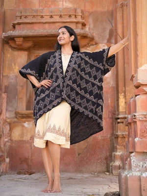 A woman poses confidently on stone steps against an ornate building, smiling upward. She wears a cream hand block-printed geometric dress with the black Cocoon Shrug draped over her shoulders, standing barefoot in style.