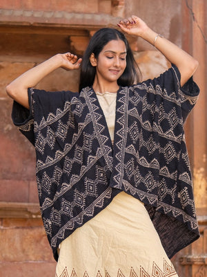 A woman stands smiling with her eyes closed, arms raised, wearing a cream cotton dress and the Cocoon Shrug—a loose black hand block-printed shawl with geometric patterns—in front of a rustic brown wall.