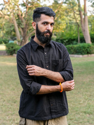 A bearded man with a mustache stands outdoors in a park, wearing the Bhadra Shirt in black paired with beige pants. Crafted from premium fabric, the Bhadra Shirt adds a refined touch to his look as he crosses his arms and gazes at the camera, surrounded by trees and grass.