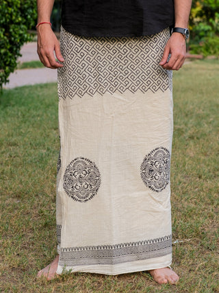 A person stands barefoot on grass, wearing an Aastha Dhoti in cream cotton with black geometric and circular patterns, their hands relaxed at their sides.