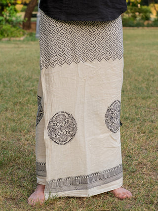 A person stands barefoot on grass, wearing the cream cotton Aastha Dhoti, featuring black Rajasthani handblock prints—geometric and floral patterns on top and circular floral motifs below.