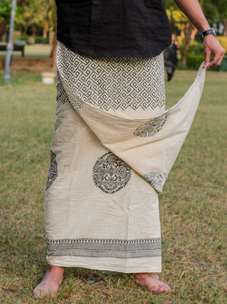 A person stands barefoot on grass, wearing a black top and the Aastha Dhoti—a soft cotton, cream-colored wrap with Rajasthani handblock print—holding one end out in a park for an effortless bohemian look.