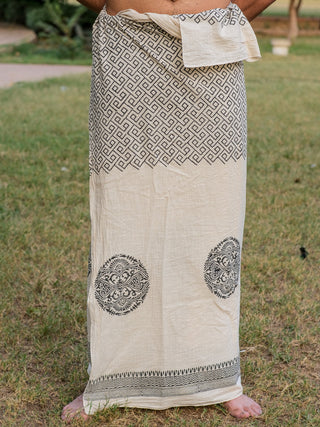 A barefoot person stands on grass, wearing the natural-colored Aastha Dhoti with black geometric and circular Rajasthani handblock prints from waist to ankles; upper body is not visible.