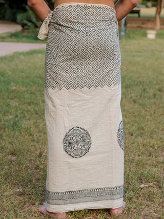 A person stands barefoot on grass, wearing the Aastha Dhoti—a cream cotton sarong featuring black geometric and circular Rajasthani handblock prints. Their upper body is not visible in the image.