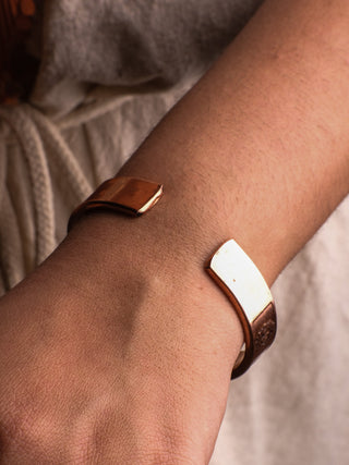 A close-up showcases a persons wrist adorned with the shariq Seven Chakra bracelet, featuring a minimalist copper-toned design. The rectangular open ends bring elegance to the light-colored fabric ensemble, harmoniously complementing and supporting ones spiritual journey.