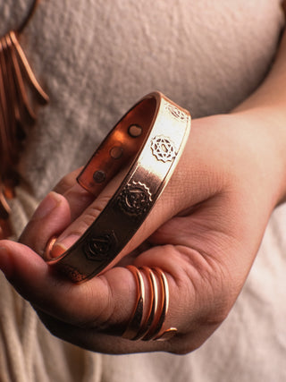 A hand gently holds a shariq Seven Chakra Bracelet, embodying a spiritual journey. The person also wears copper rings, enhancing their energy centers against a soft beige fabric background.