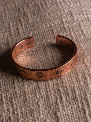 A rustic copper bracelet by shariq, featuring engraved geometric patterns, rests on textured beige fabric. Reminiscent of the Seven Chakra Bracelet, it includes small circular magnets inside—ideal for a spiritual journey.