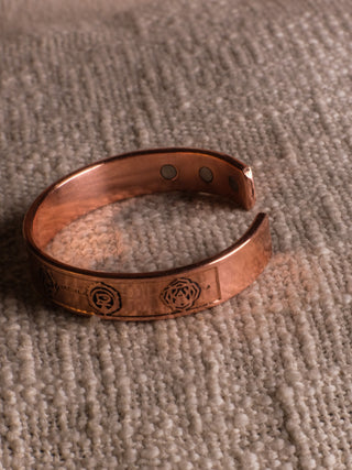 A Seven Chakra Bracelet by shariq, featuring engraved designs and circular magnets on the inside, lies gracefully on a textured beige fabric surface.