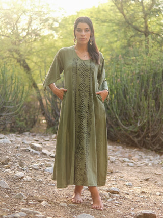A barefoot woman stands outdoors on a rocky path, wearing the Garga Dress—an elegant olive green dress with block printing and geometric patterns. Sunlit green trees and shrubs fill the background.