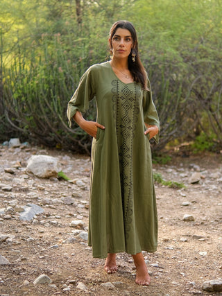 A woman with long dark hair stands barefoot outdoors on a rocky path, surrounded by greenery. She wears the elegant Garga Dress in green with geometric block prints, looking ahead with her hands in her pockets.