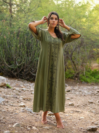 A woman stands barefoot on a rocky path amid greenery, wearing the olive-green Garga Dress with black block print. She gazes upward, touching her ears, surrounded by plants and trees.