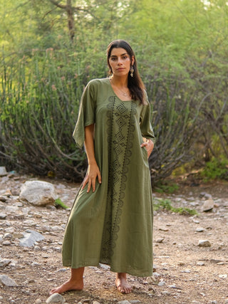 A woman with long dark hair stands barefoot outdoors on rocky ground, wearing the Garga Dress in olive green with geometric block print, surrounded by lush trees and foliage.